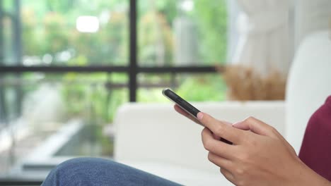 Schöne-asiatische-Frau-Zeit-genießen-und-sitzen-auf-dem-modernen-Sofa-vor-Fenster-Entspannung-in-ihrem-Wohnzimmer-mit-Smartphone-zum-sprechen,-lesen-und-SMS.-Lebensstil-Asia-Frau-am-Wohnkonzept.