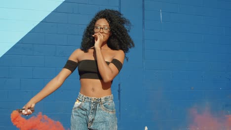 Beautiful-young-woman-holding-colorful-smoke-grenade-dancing-outside-against-blue-wall