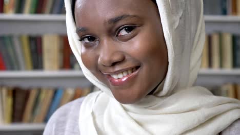 Cara-de-niña-musulmana-africana-encantadora-joven-en-hijab-está-mirando-a-cámara,-religioun-concepto,-booksheves-en-el-fondo