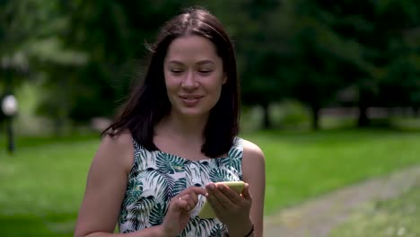 Close-up-of-a-happy-looking-asian-woman-walking-in-the-park-and-typing-sms-on-the-yellow-smartphone.