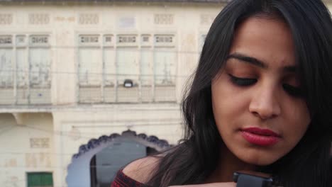 Young-attractive-woman-busy-with-smart-watch-talks-listens-communicates-smiles-laughs-happy-joy-looks-at-the-camera-with-a-traditional-local-Rajasthani-house-facade-in-the-backdrop-handheld-mid-shot