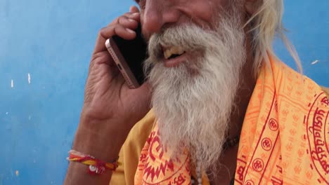 Tilt-up-to-Sadhu,-Hindu-priest,-talking-on-smart-phone-with-a-blue-background