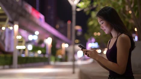 Schöne-asiatische-Frau-draußen-in-der-Nacht-mit-Handy