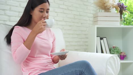 Mujer-asiática-joven-alegre-beber-café-caliente-o-té-disfrutando-mientras-está-sentado-en-su-sala-de-estar-en-casa.-Atractiva-mujer-asiática-feliz-sosteniendo-una-taza-de-café.