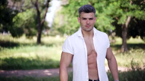 Hombre-guapo-joven-en-camisa-blanca-sonriendo,-mirando-a-cámara-en-el-parque-en-un-caliente-día-de-verano.-Retrato-de-joven-sonriente-deportista-en-el-Parque