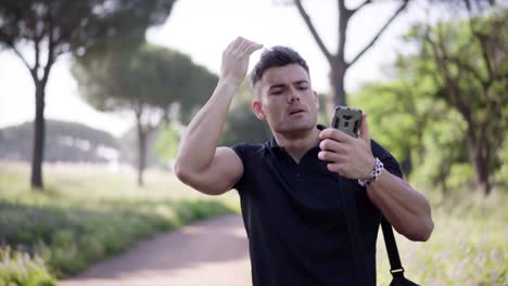 Retrato-de-un-joven-deportista-guapo.-Un-hombre-hermoso-deportivo-manos-su-pelo-mirando-el-teléfono-en-el-parque-de-la-ciudad-en-el-verano