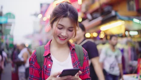 Asiatische-Frau-touristischen-Backpacker-lächelnd-und-mit-Smartphone-alleine-Urlaub-im-freien-unterwegs-Stadtstraße-in-der-Khao-San-Road-in-Bangkok-Bangkok---Thailand.