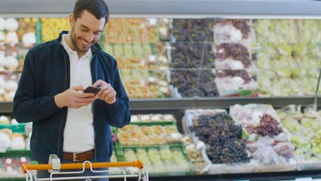 En-el-supermercado:-hombre-guapo-utiliza-teléfono-inteligente-mientras-que-en-la-sección-de-producto-fresco-de-la-tienda.-Hombre-inmerso-en-la-navegación-por-Internet-en-su-teléfono-móvil-en-el-fondo-coloridas-frutas-y-verduras-orgánicas.