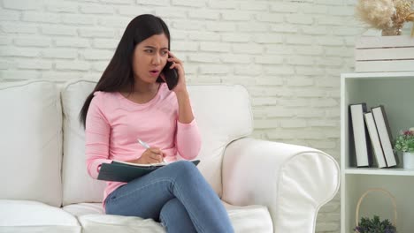 Joven-atractiva-mujer-asiática-escribir-portátil-y-smartphone-que-habla-en-el-sofá-en-la-acogedora-sala-de-estar-en-casa.-Estilo-de-vida-casual-en-el-hogar.