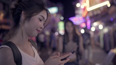 Backpacker-de-turismo-asiático-mujer-sonriendo-y-con-smartphone-viajando-solo-vacaciones-al-aire-libre-por-la-ciudad-calle-de-Khao-San-road-en-Bangkok-Bangkok---Tailandia.