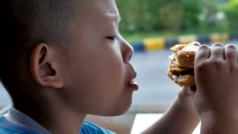 Cerca-niño-asiáticos-comer-hamburguesa,-lindo-muchacho-feliz-con-hamburguesa-en-el-restaurante.-Video-Slow-motion