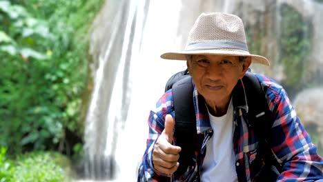 Mano-de-Asia-de-hombre-Senior-con-pulgar-arriba-sentado-en-frente-de-la-cascada.-Éxito,-vida-aventuras-de-viajes-y-turismo-con-mochila.-Video-Slow-motion