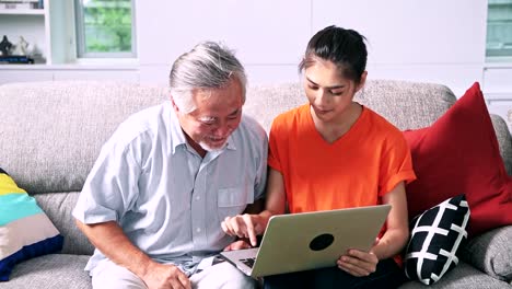 Tochter-Unterricht-ihres-Vaters-Computerkenntnisse-im-Wohnzimmer.-Asiatischer-Mann-mit-weißem-Bart-und-junge-Frau-sitzt-im-Wohnzimmer-mit-Laptop.-Familie-Senior-Lifestyle-Konzept.