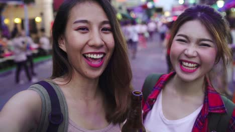 Viajero-mochilero-blogger-par-de-lesbianas-lgbt-de-Asia-las-mujeres-viajar-con-smartphone-para-selfie-en-Tailandia.-Mujer-bebiendo-alcohol-o-cerveza-en-Khao-San-Road,-la-calle-más-famosa-de-Bangkok.