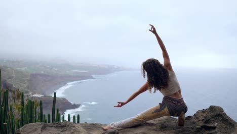 A-woman-sitting-on-the-edge-of-a-cliff-in-a-pose-war-overlooking-the-ocean-raise-her-hands-up-and-inhale-the-sea-air-while-doing-yoga