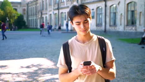 Concentrated-asian-boy-is-typing-on-his-phone-and-thinking-about-answer-while-standing-on-the-road-near-buildings,-sunny-mood