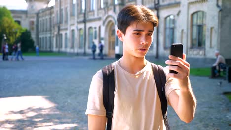 Enjoyable-asian-man-is-having-phone-conversation,-web-camera-using,-standing-in-front-of-the-university-building,-sunny-day