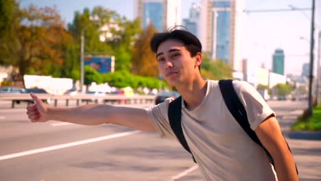 Sweet-asian-boy-is-catching-the-car-on-the-road,-smiling-and-showing-braces-while-standing-on-highway-alone,-perfect-weather-for-travelling