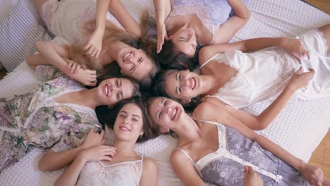 Pyjama-Party,-sechs-junge-Frauen-in-stilvollen-sexy-Nachtwäsche-lächelnd-auf-dem-Bett-liegen-und-schauen-Sie-direkt-in-der-Kamera-im-Raum