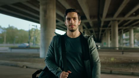 Portrait-shot-of-an-Athletic-Young-Man-Walking-From-the-Road-Under-a-Bridge-in-an-Urban-Environment.-He's-Wearing-a-Grey-Hoodie-and-a-Sports-Bag.