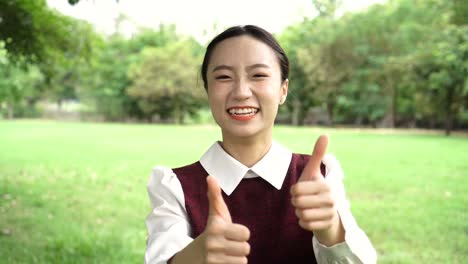 Joven-atractiva-mujer-asiática-en-uniforme-mostrando-los-pulgares-para-arriba-gesto-en-el-parque-al-aire-libre,-como-gesticular-de-la-escuela