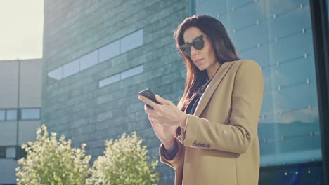 Elegant-Businesswoman-Uses-Smartphone-to-Conduct-Business-while-Standing-Near-Modern-Glass-Building.-Beautiful-Stylish-Woman-Wearing-Coat-and-Dark-Glasses-Walks-in-Modern-City-Urban-Environment.