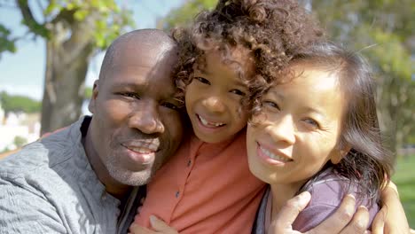 Multiethnic-family-hugging-curly-daughter-in-park