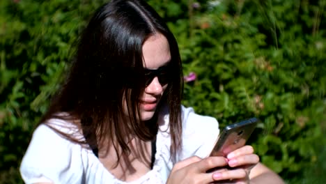 Woman-is-typing-a-message-on-mobile-phone-sitting-in-park-in-sunny-day,-chatting.