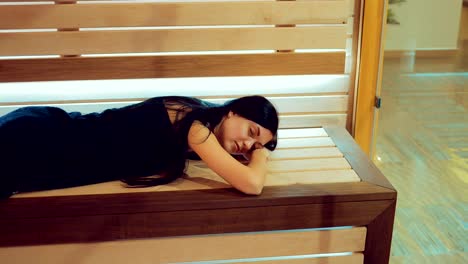 Beautiful-brunette-girl-is-lying-on-a-wooden-bench-in-a-sauna-behind-a-glass-door.-Girl-is-resting-with-her-eyes-closed.-Span-camera