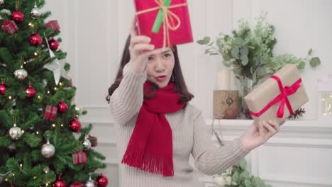 Cheerful-happy-young-Asian-woman-holding-christmas-gifts-smiling-to-camera-in-her-living-room-at-home-in-Christmas-Festival.-Lifestyle-woman-celebrate-Christmas-and-New-year-concept.