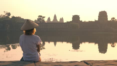 Woman-visiting-Angkor-Wat-ruins-at-sunrise,-famous-travel-destination-Cambodia.-Reflection-on-water-pond-and-sunburst,-soft-light,-toned.
