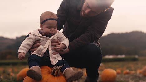 A-baby-sitting-on-a-pumpkin-at-a-pumpkin-patch,-with-her-dad-holding-her-up