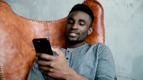 Portrait-of-young-African-man-sitting-in-the-chair,-using-Smartphone.-A-handsome-male-smiling-and-talking-on-the-phone