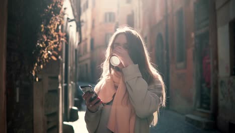 Portrait-of-young-happy-woman-walking-in-city-and-using-smartphone.-Girl-browse-the-Internet-and-drinking-coffee