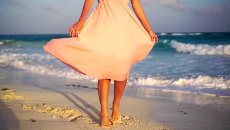 Schöne-junge-Frau-im-Sommerurlaub-am-weißen-Strand.-Nahaufnahme-weibliche-Beine-am-Strand-im-Sonnenuntergang