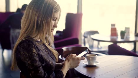 Joven-muy-femenina-con-teléfono.-Joven-mujer-atractiva-elegante-vestido-sentado-en-la-mesa-con-café-y-uso-de-smartphone