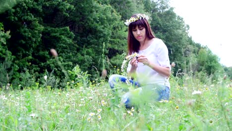 Verano,-en-medio-de-un-césped-de-la-manzanilla,-en-un-bosque,-una-mujer-joven,-una-morena-teje-una-guirnalda-de-chamomiles