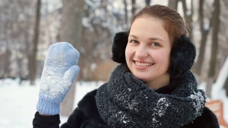 Pretty-lady-waves-her-hands-in-mittens-to-the-camera-at-winter-park-background