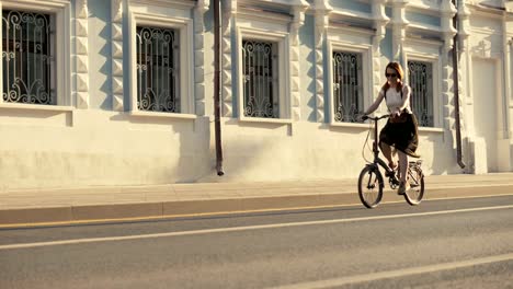 Rote-kurzhaarige-Frau-Reiten-Fahrrad-in-der-Stadt-unterwegs-Hintergrund-Gebäude