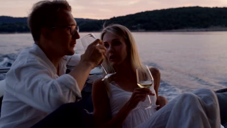 Young-Couple-Talk,-Drink-Champagne-in-the-Stern-of-the-Moving-Yacht.-They-Have-Great-Romantic-Evening.-In-the-Background-Island-with-Small-Village.