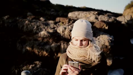 Portrait-of-young-blonde-woman-standing-in-mountains-in-sunny-day-and-taking-selfie-photo-on-smartphone