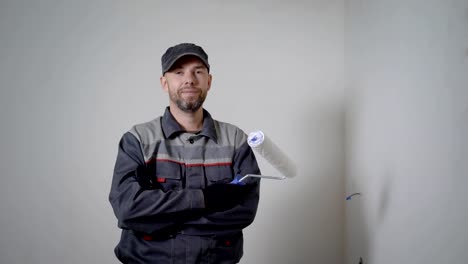 cheerful-repairman-is-standing-in-apartment,-holding-paint-roller,-crossing-hands-in-background-of-white-empty-walls