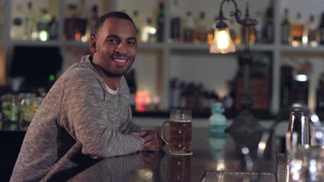 Toma-de-retrato-de-un-hombre-afroamericano-atractivo-joven-sonriendo-y-tomando-un-sorbo-de-su-cerveza-en-un-bar