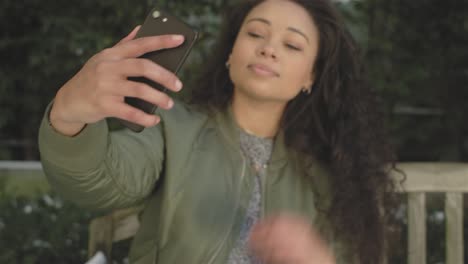 Porträtaufnahme-von-eine-Beautirul-junge-Frau,-die-eine-Selfie-auf-ihrem-Handy-auf-eine-hölzerne-Parkbank-sitzend