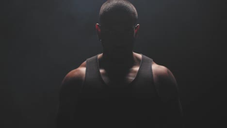 Portrait-shot-of-a-muscular-black-man-staring-at-the-camera-on-a-foggy-dark-background.