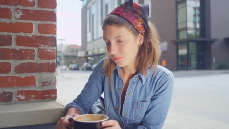 Hipster-young-woman-sitting-outside-a-coffee-shop-smiles-and-drinks-her-coffee,-portrait