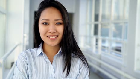 Portrait-of-young-beautiful-pretty-female-student-of-asian-ethnicity-standing-in-wide-white-hallway-indoors-looking-at-camera-and-smiling-positively