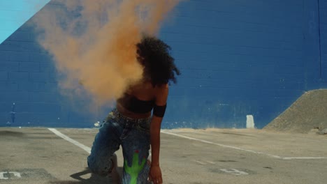 Beautiful-young-woman-holding-colorful-smoke-grenade-dancing-outside-against-blue-wall