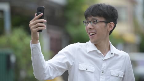 Young-happy-Asian-businessman-video-calling-in-the-streets-outdoors