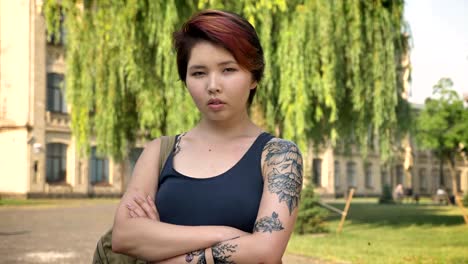 Portrait-of-young-asian-women-with-tattoo-standing-with-crossed-hands-and-looking-in-camera-in-park-near-university,-confident-and-serious